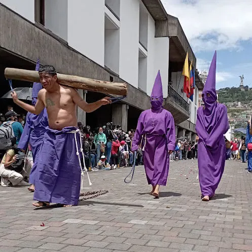 Penitente descalzo arrastrando un madero pesado por el empedrado, con otros cucuruchos detrás