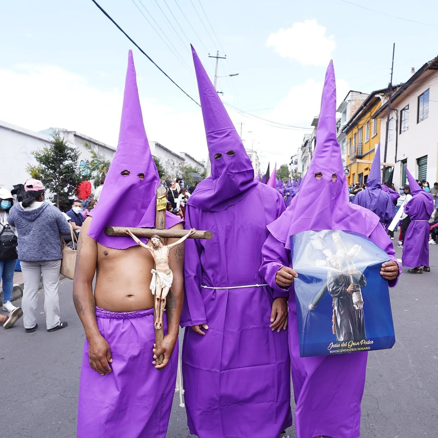Tres cucuruchos en la calle; uno carga una cruz de madera, otro sostiene una lámina de Jesús del Gran Poder