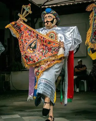 Danzante de Yaruquíes en plena danza, con una pierna en alto, sosteniendo una cruz dorada y el pañolón bordado