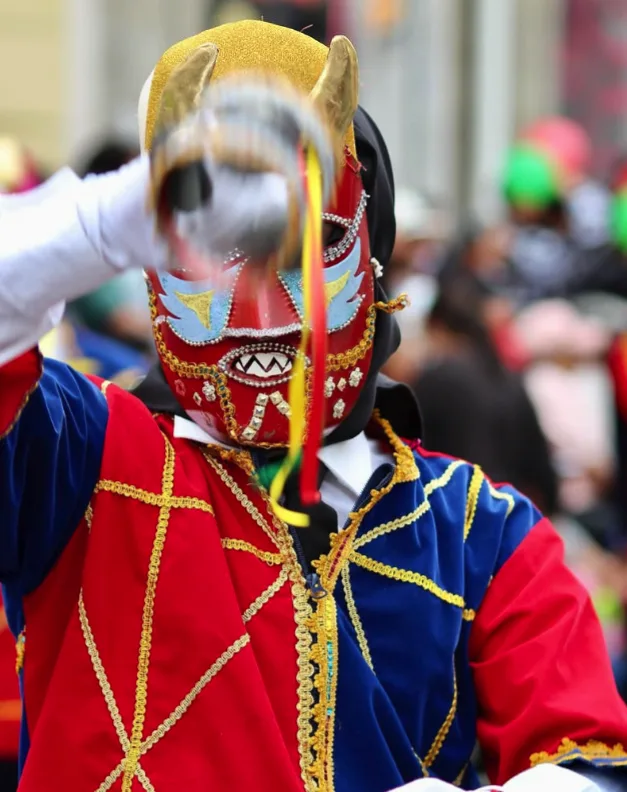 Primer plano de máscara roja con cuernos dorados y dientes plateados, sobre traje rojo y azul con galón dorado